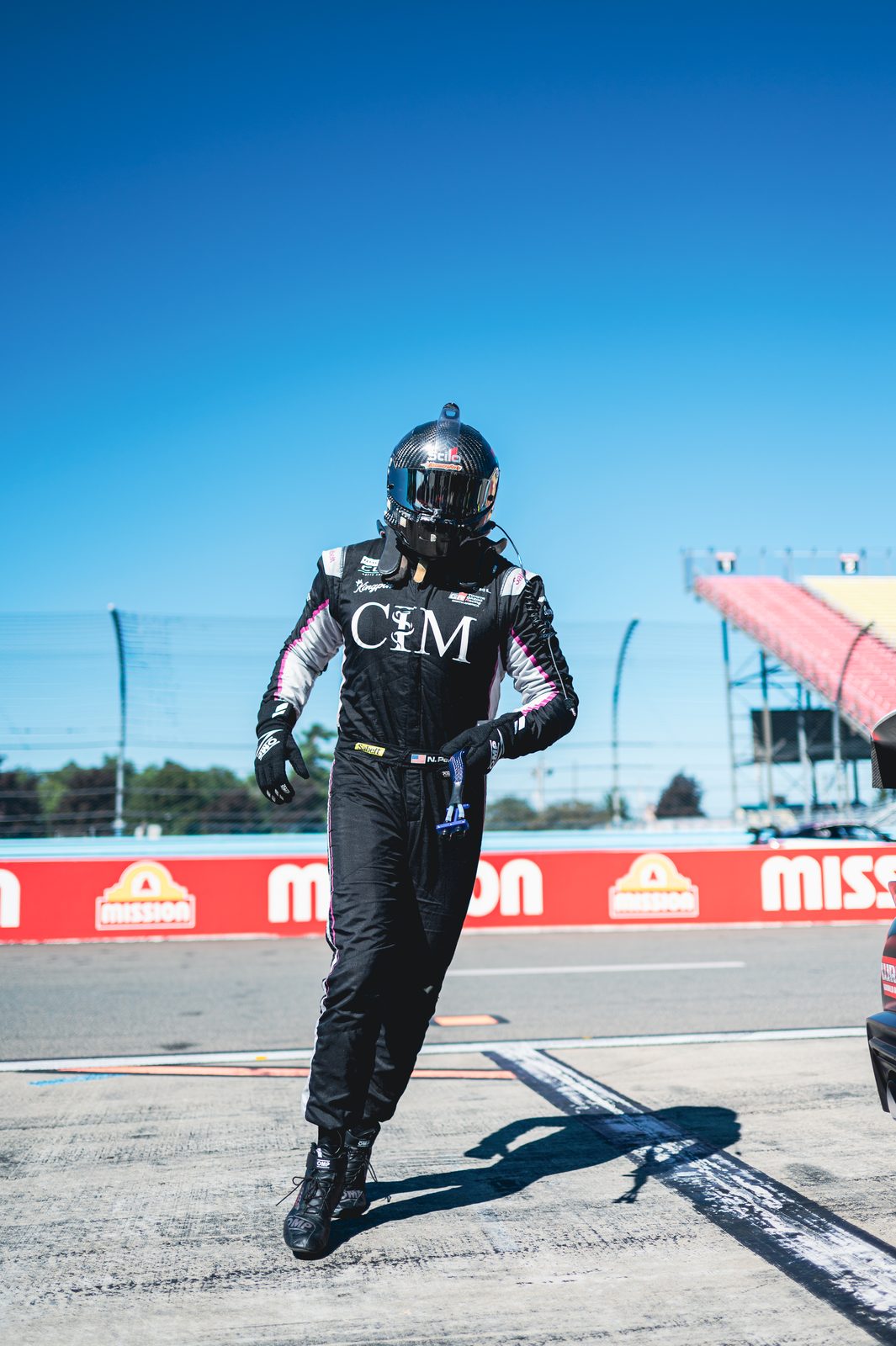 Driver in a black race suit and helmet walking across the pit lane